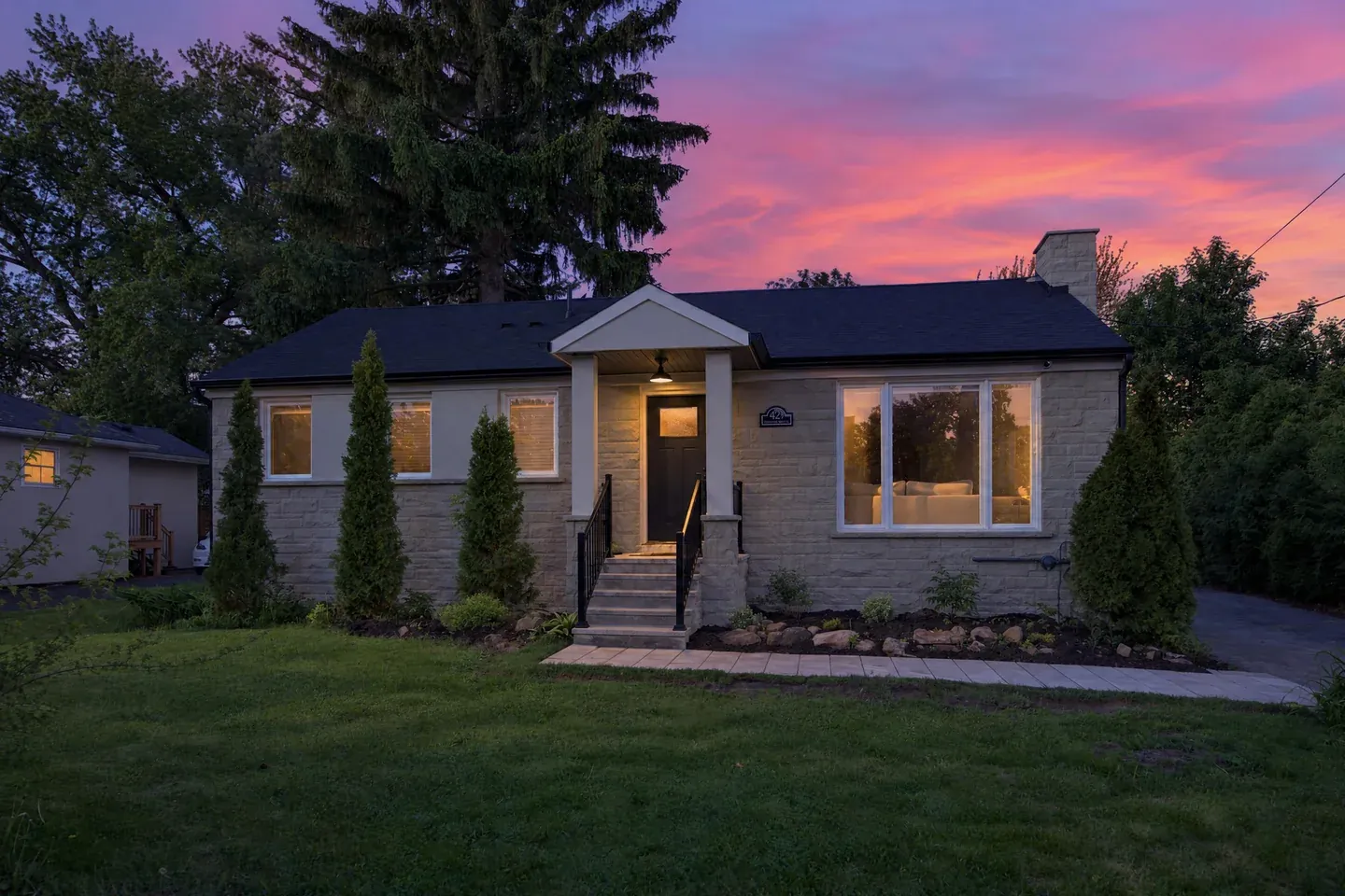 The same bungalow exterior edited with AI to dusk: glowing windows and porch light under a pink and purple sunset sky